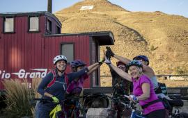 A group of adult bicyclists give each other a high-five in front of the historic train caboose at the base of S Mountain